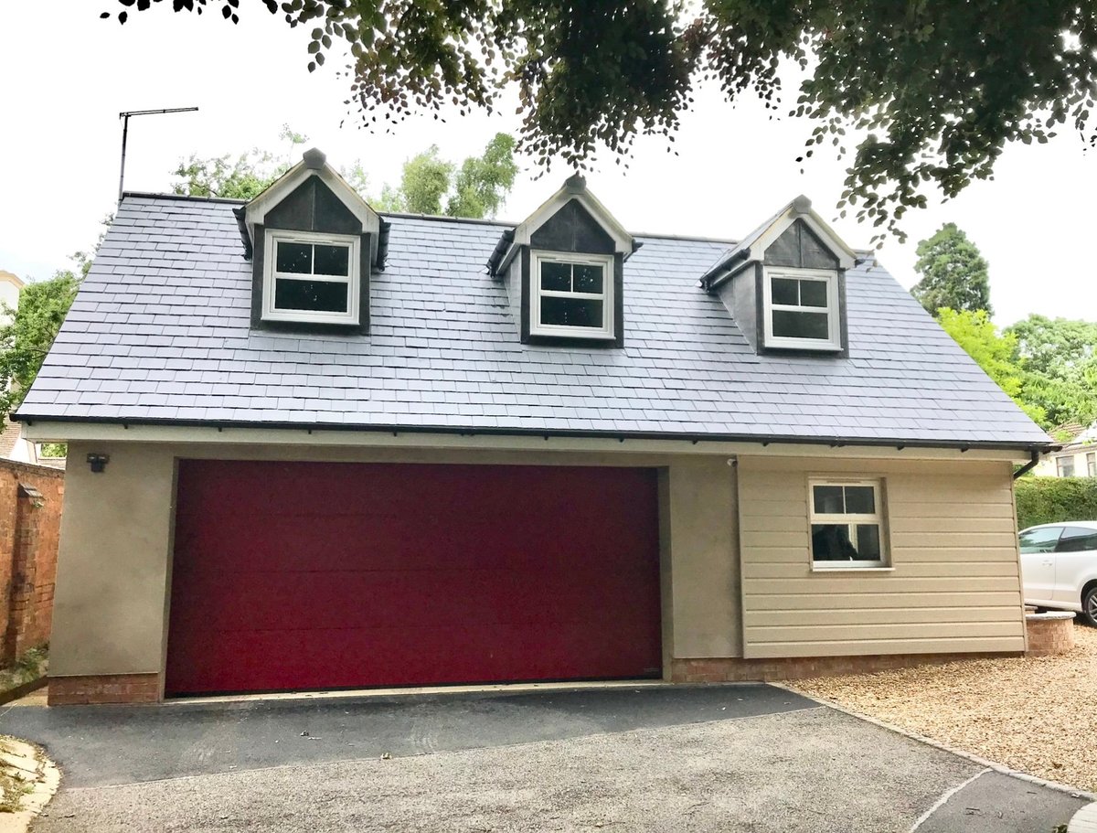 Modern garage with burgundy double doors, slate roof with three dormer windows, cream siding, and gravel driveway