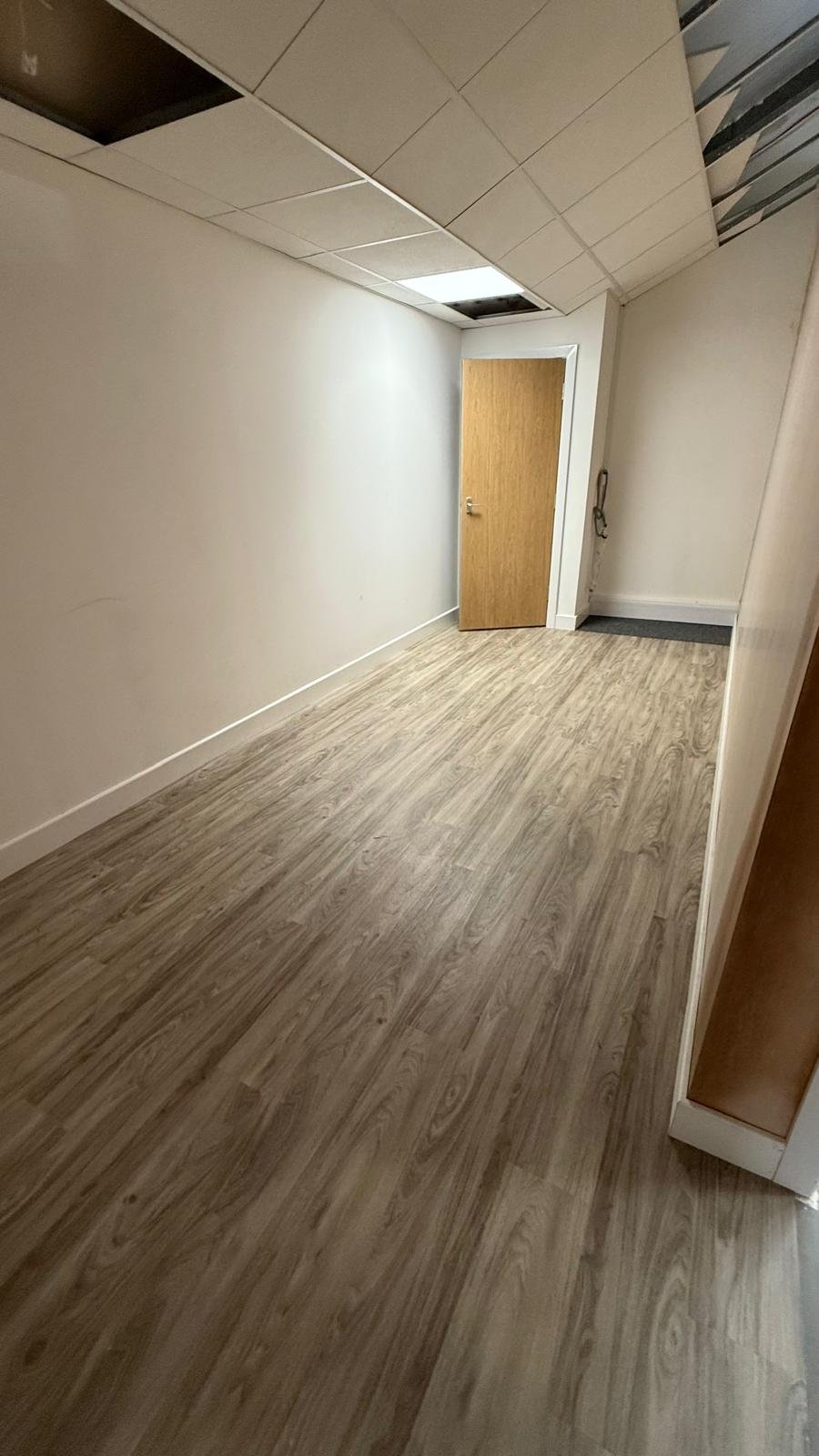 Empty office or commercial space with light gray walls, drop ceiling, wood-textured vinyl flooring, and a wooden door