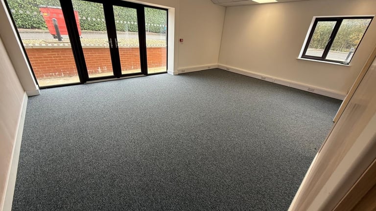 Empty office or commercial space with gray carpet flooring, white walls, and large black-framed windows and glass doors overlooking a brick building