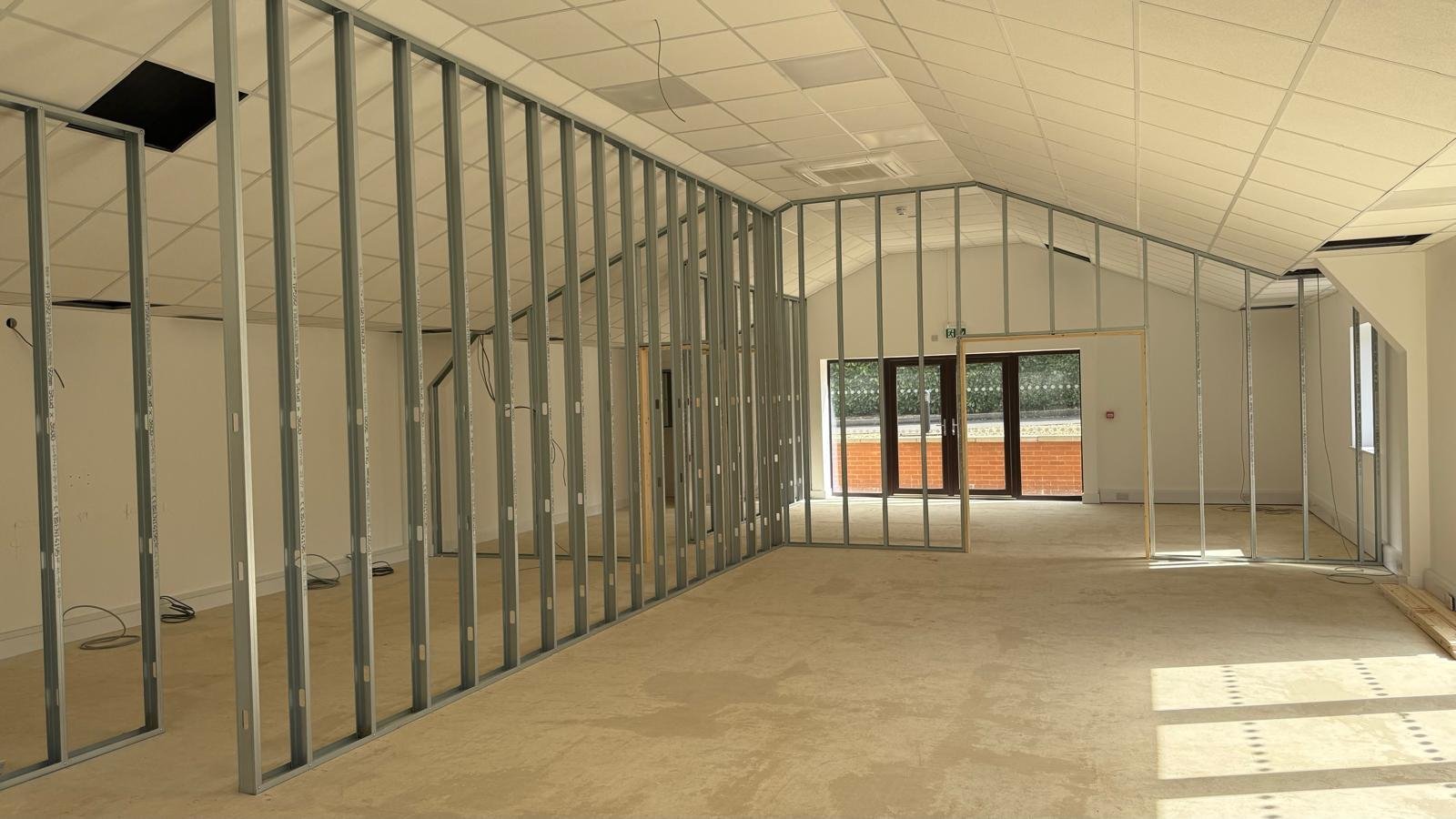 Empty office or retail space with metal framing and drywall under construction, featuring a tiled ceiling and large windows