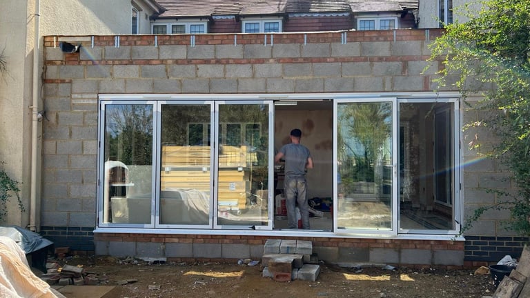 Construction site showing large glass sliding doors and windows being installed on a brick residential building with a worker visible inside