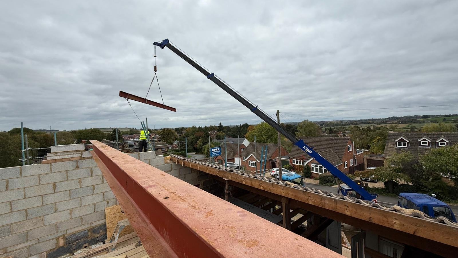 Construction worker using crane to lift red beam on residential building site with suburban landscape in background.