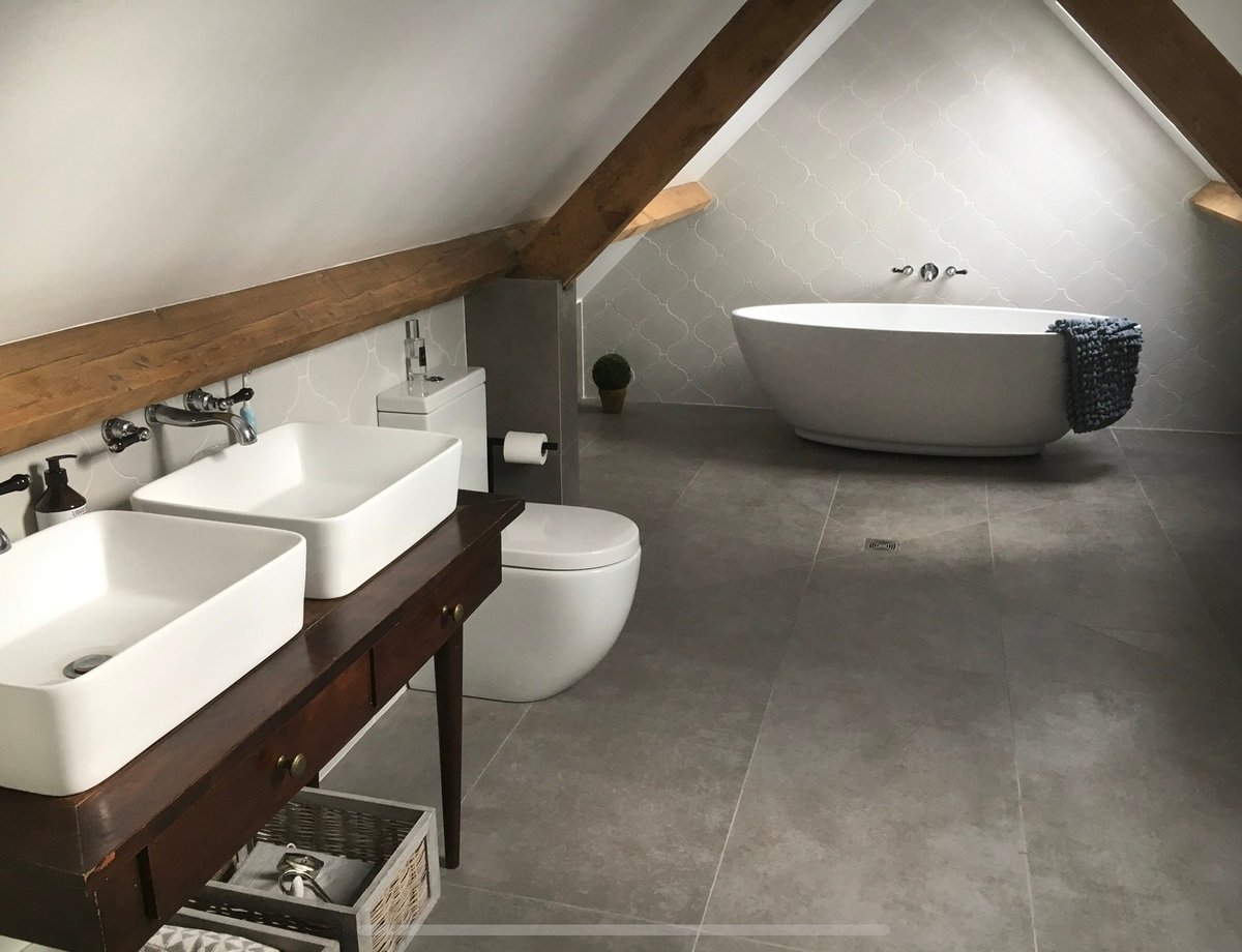 Modern attic bathroom with dual sinks, standalone tub, toilet, and gray tile flooring under sloped wooden beams