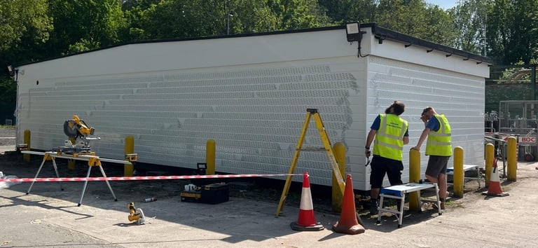Workers in safety vests installing white siding on a mobile home structure with yellow ladders and equipment