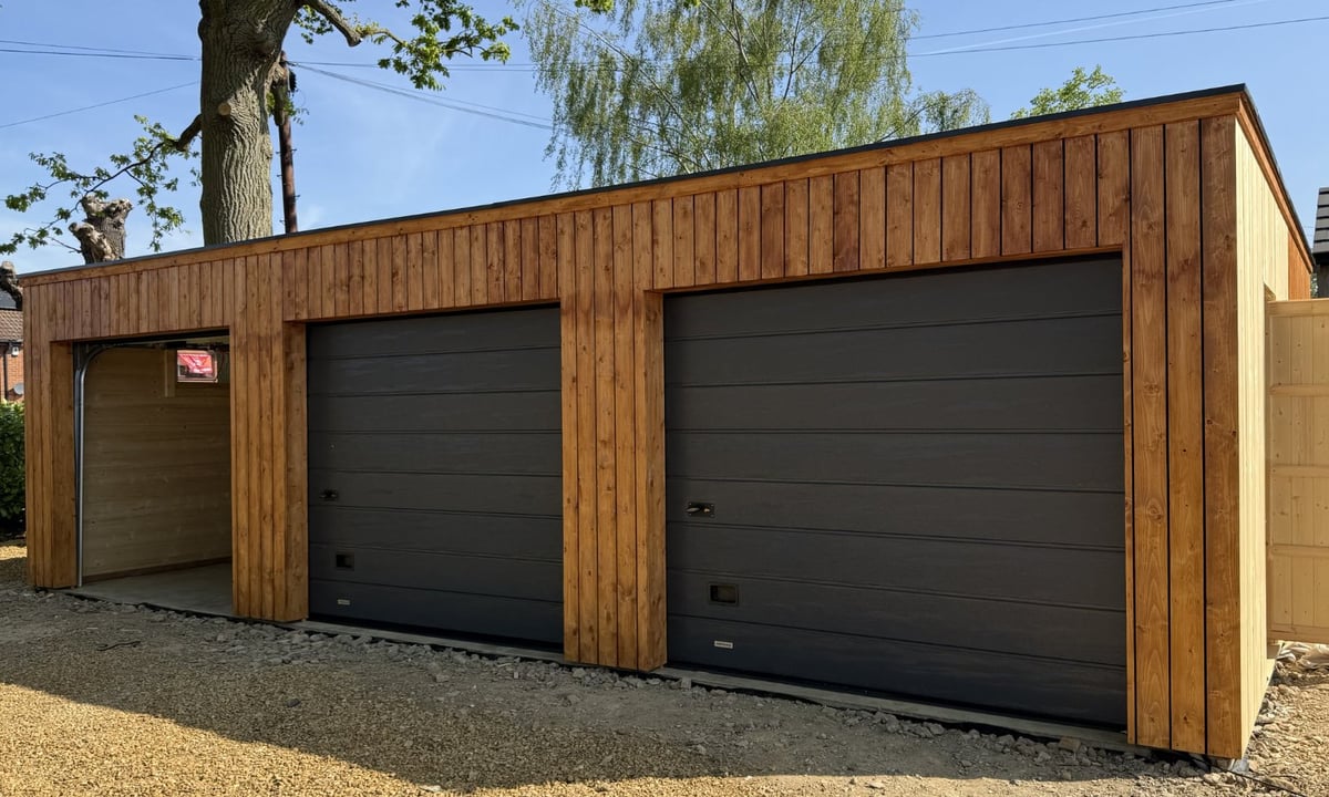 Modern wooden garage with three black metal doors and gravel driveway, surrounded by trees
